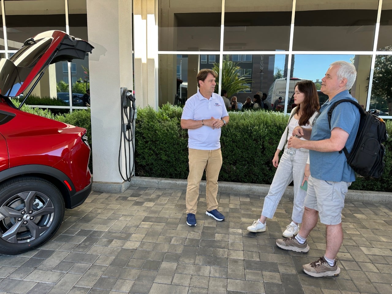 Two car shopping customers speak to a man in khaki pants and a polo shirt while looking at a new vehicle for purchase.